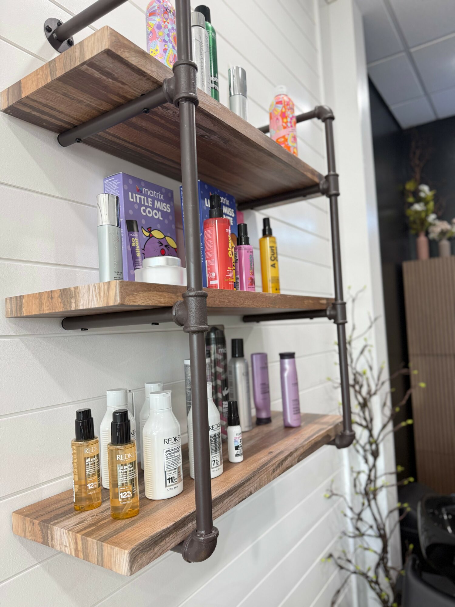 Three wooden shelves with various bottles and containers mounted on a white wall, with a plant visible in the background.