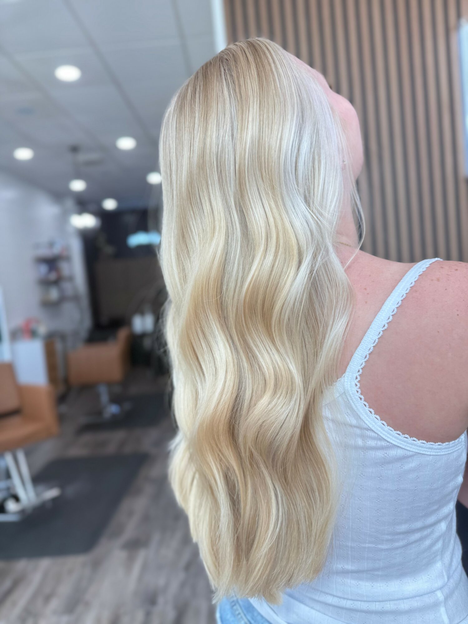 Back view of a woman with long, wavy blonde hair in an indoor setting, wearing a white top with lace straps.