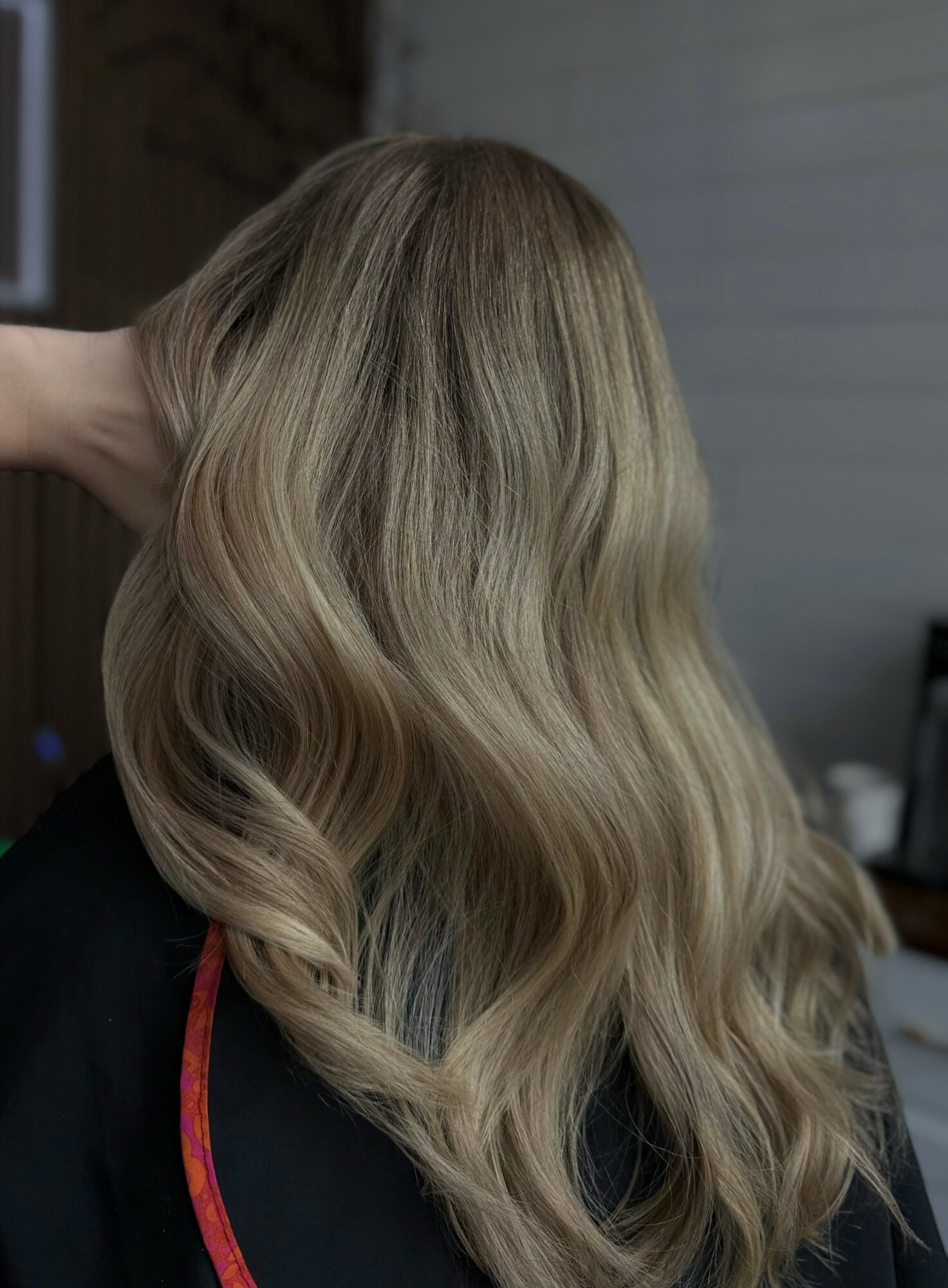 Person with long, wavy blonde hair, viewed from the back, indoors with a blurred background.