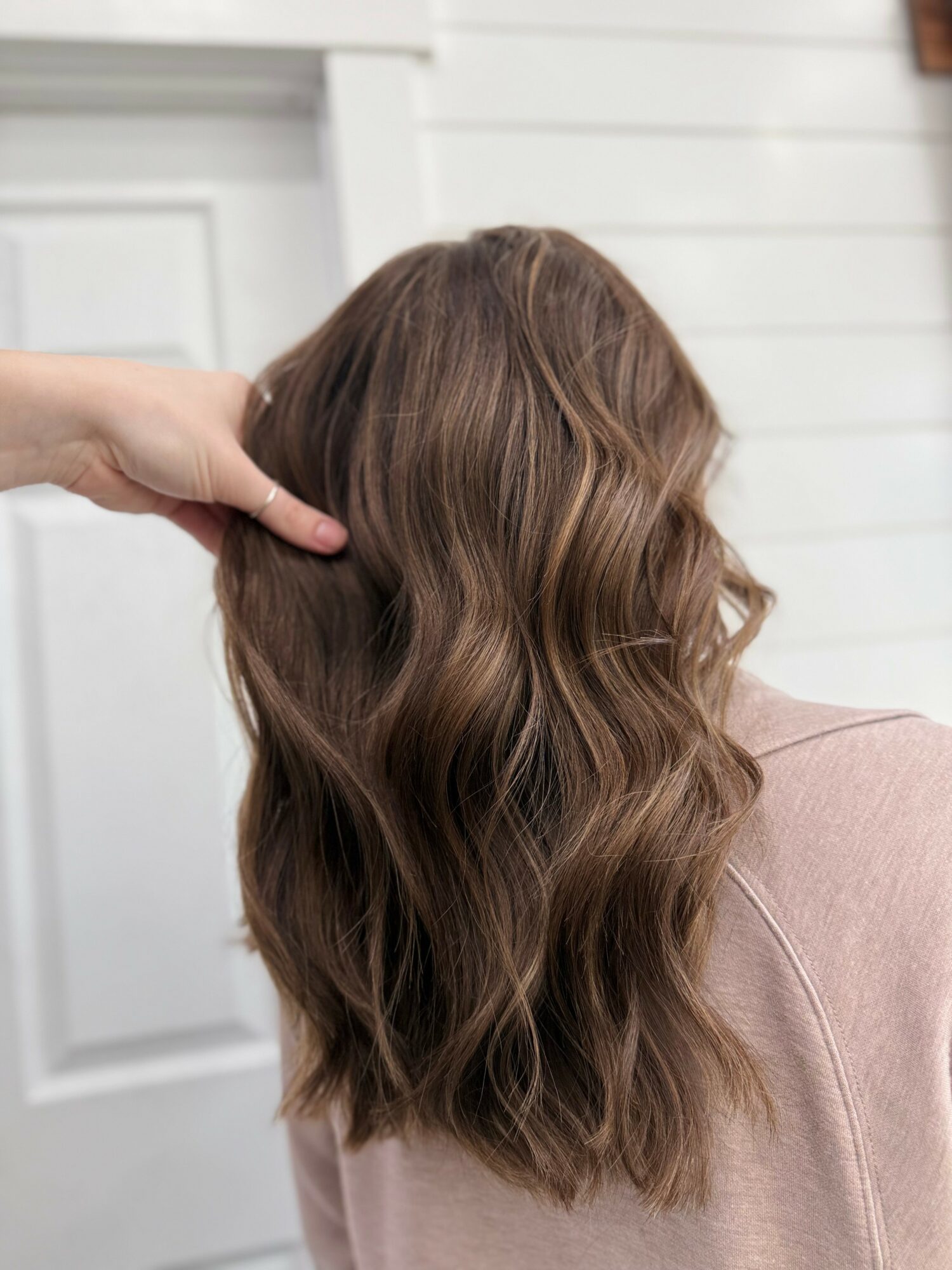 Person with wavy, brown hair viewed from behind, sitting in front of a white wall and door, with hand touching hair.