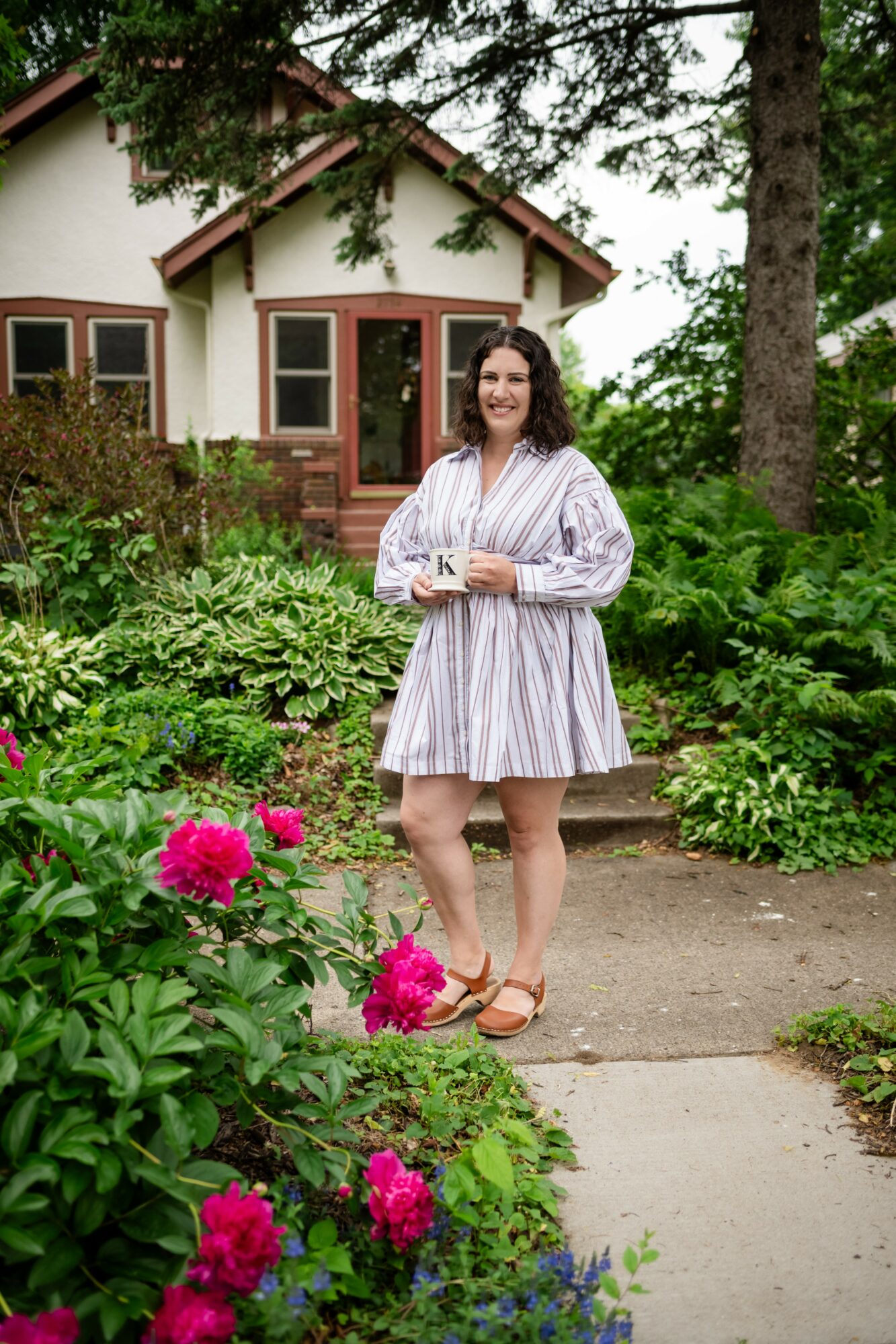Woman standing on garden path in front of house, surrounded by greenery and pink flowers, smiling, holding a cup.