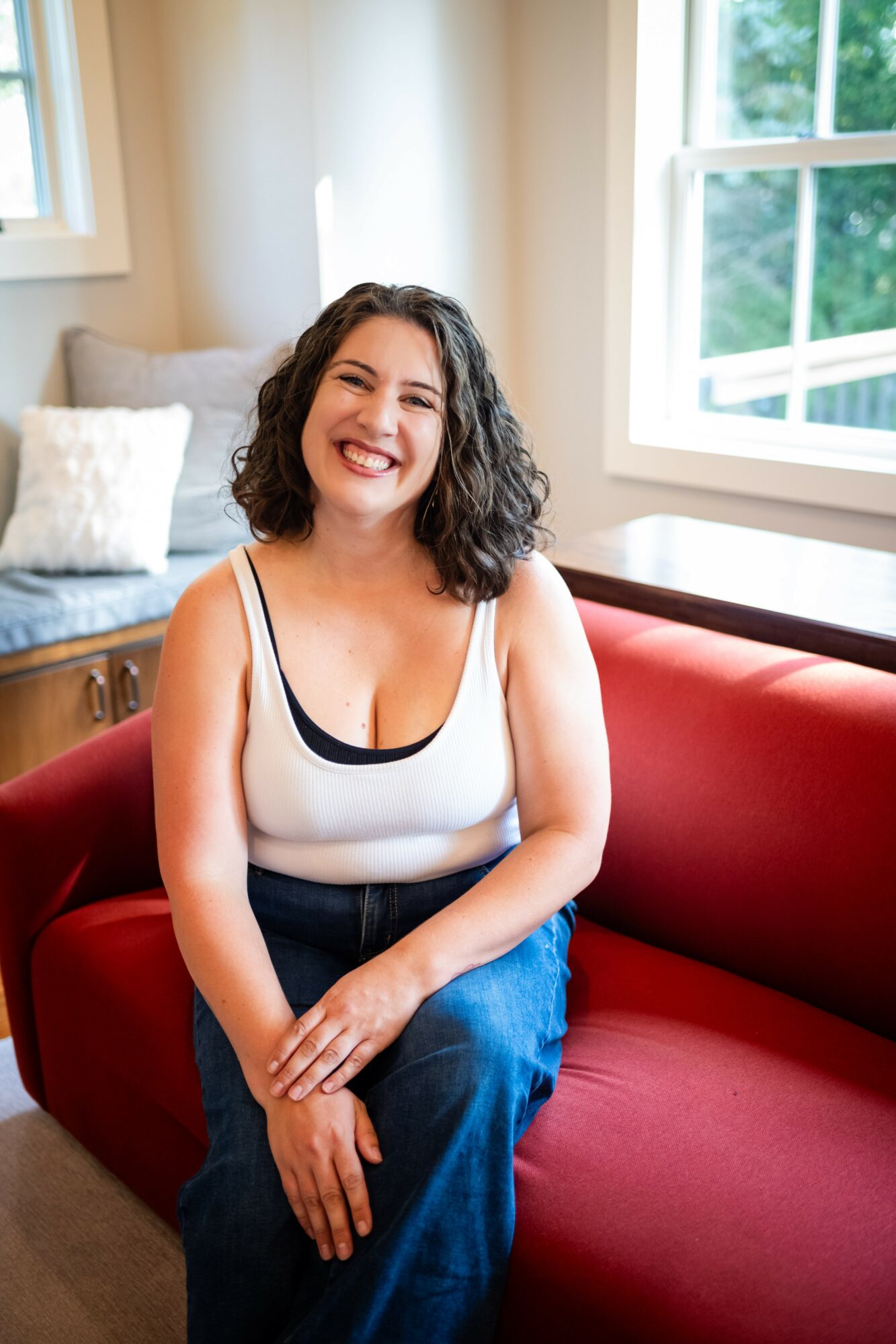 Young woman with curly hair smiling, sitting on a red couch in a bright room with windows, wearing a white tank top and jeans.