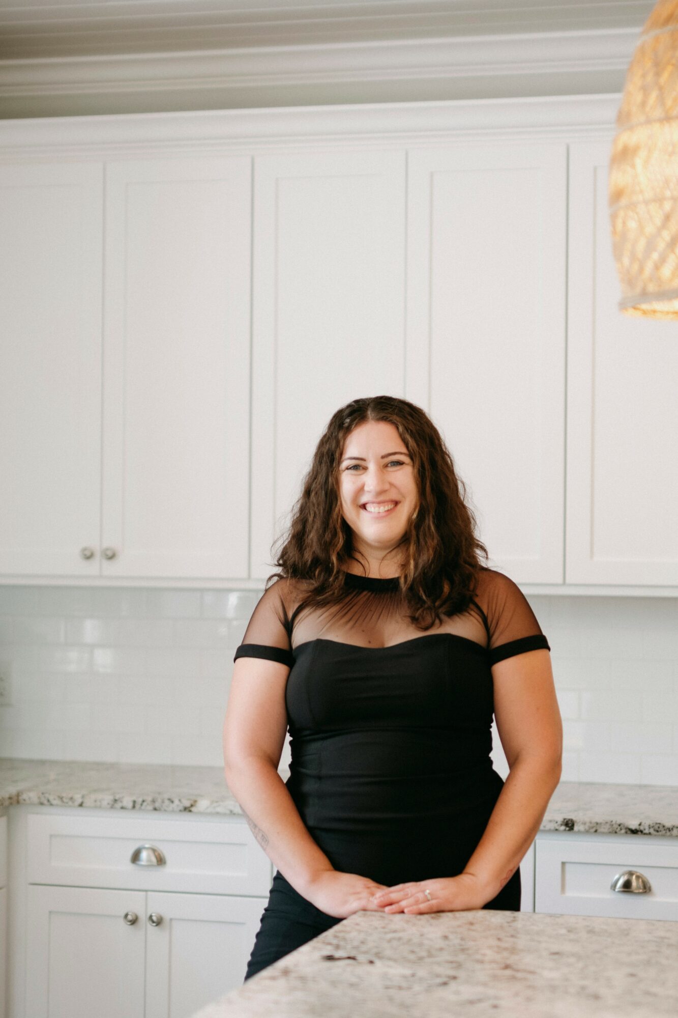Young woman with curly hair smiling, standing in a kitchen with white cabinets and marble countertop.