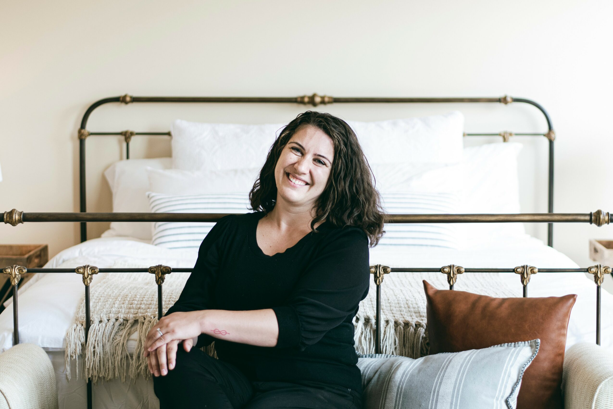 Smiling woman with dark curly hair sitting on a sofa in front of a bed with white bedding and pillows, brown and striped cushions.