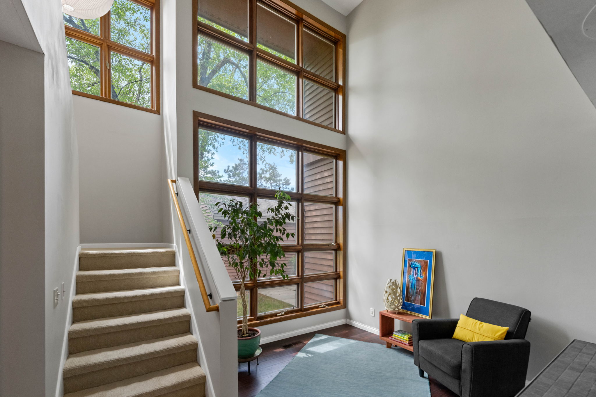 Interior of a modern home with large windows, a staircase, a potted plant, and a seating area with a black chair and yellow cushion.