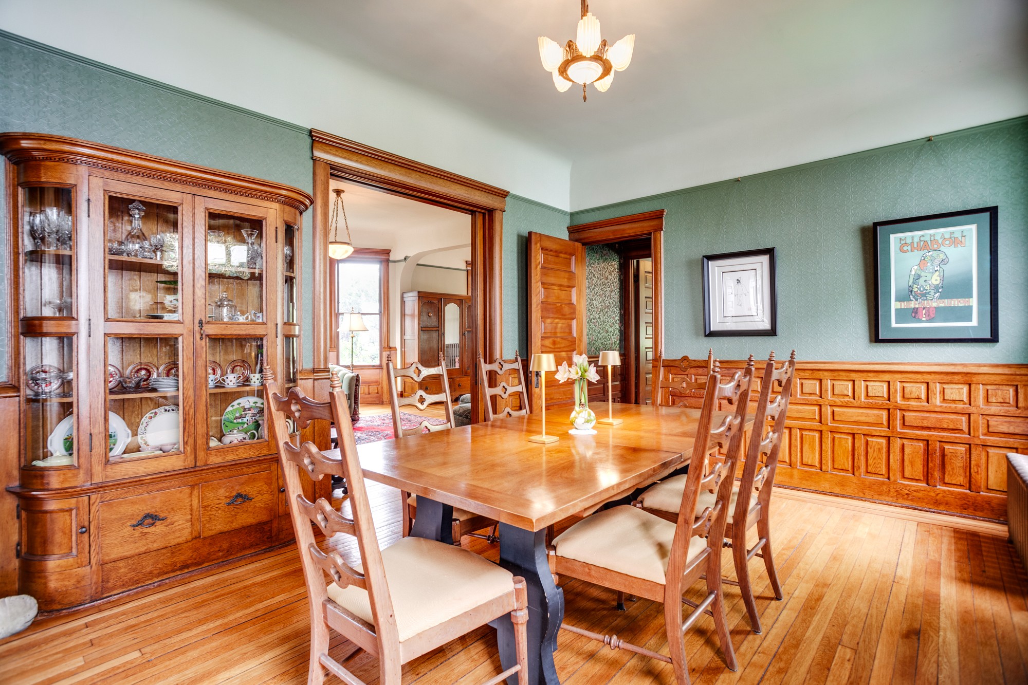 Wooden dining table with six chairs in a room with green walls, wooden trim, and framed artwork, hardwood floor, and a ceiling light.