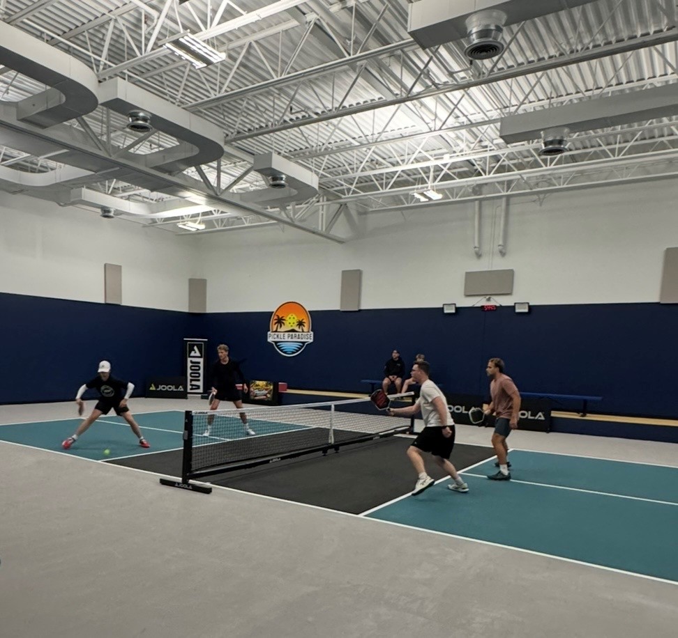People playing pickleball on an indoor court with a blue wall and ceiling, and a logo on the wall, four players visible.