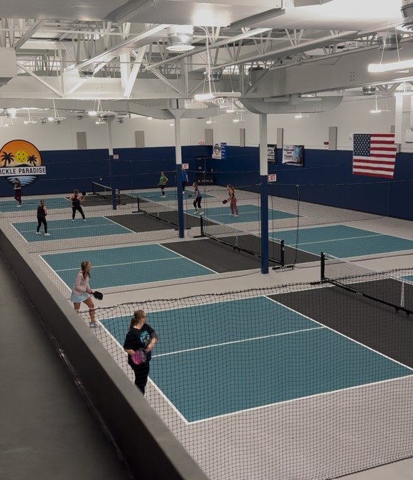 Indoor tennis courts with players, net, and American flag, ceiling with white beams, and a sunset logo on the wall.