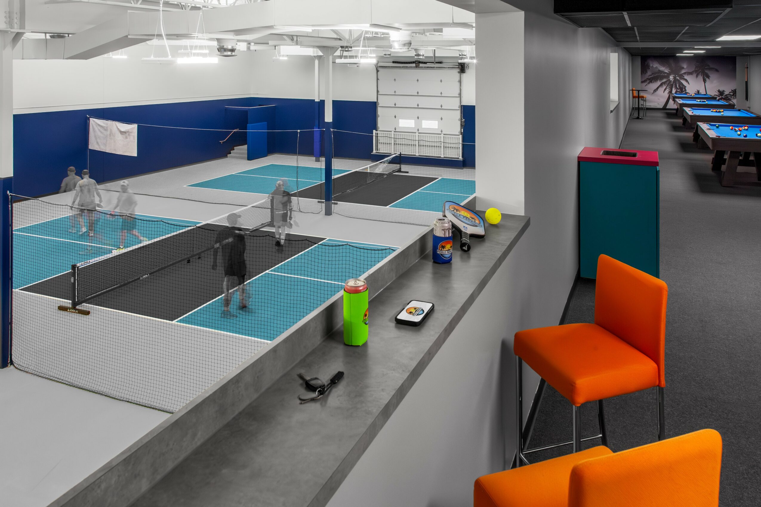 Indoor recreational area with multiple mini tennis or ping pong courts, seating, and game tables, with a person playing in the background.
