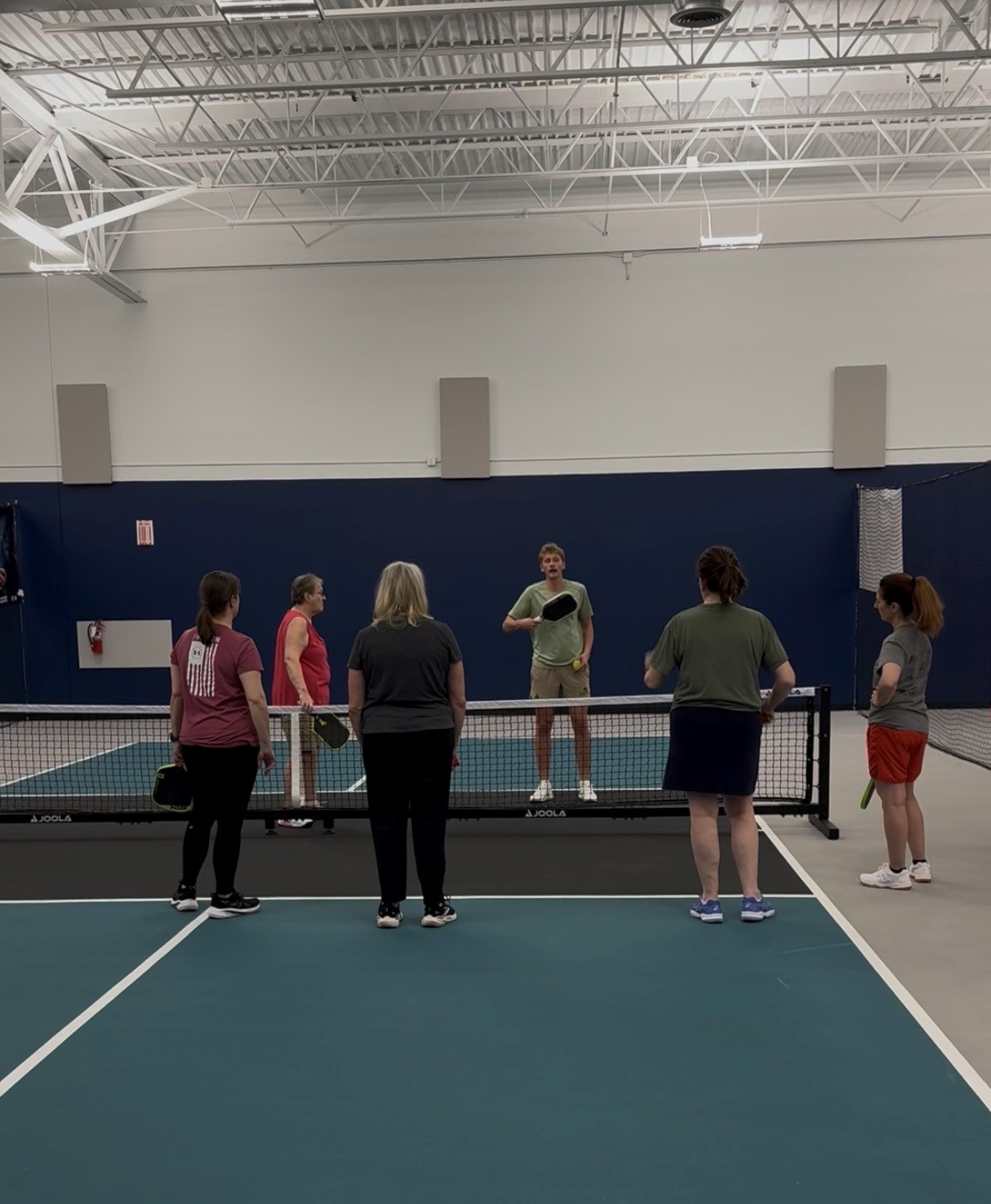 Group of six people standing around a tennis net inside a gymnasium, listening to a person holding a tennis racket.