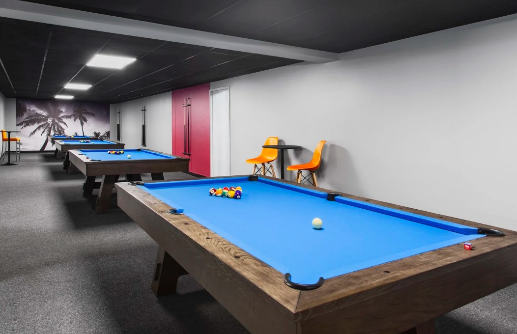 Pool table with blue felt and balls, chairs, and a mural in the background, in an indoor game room.