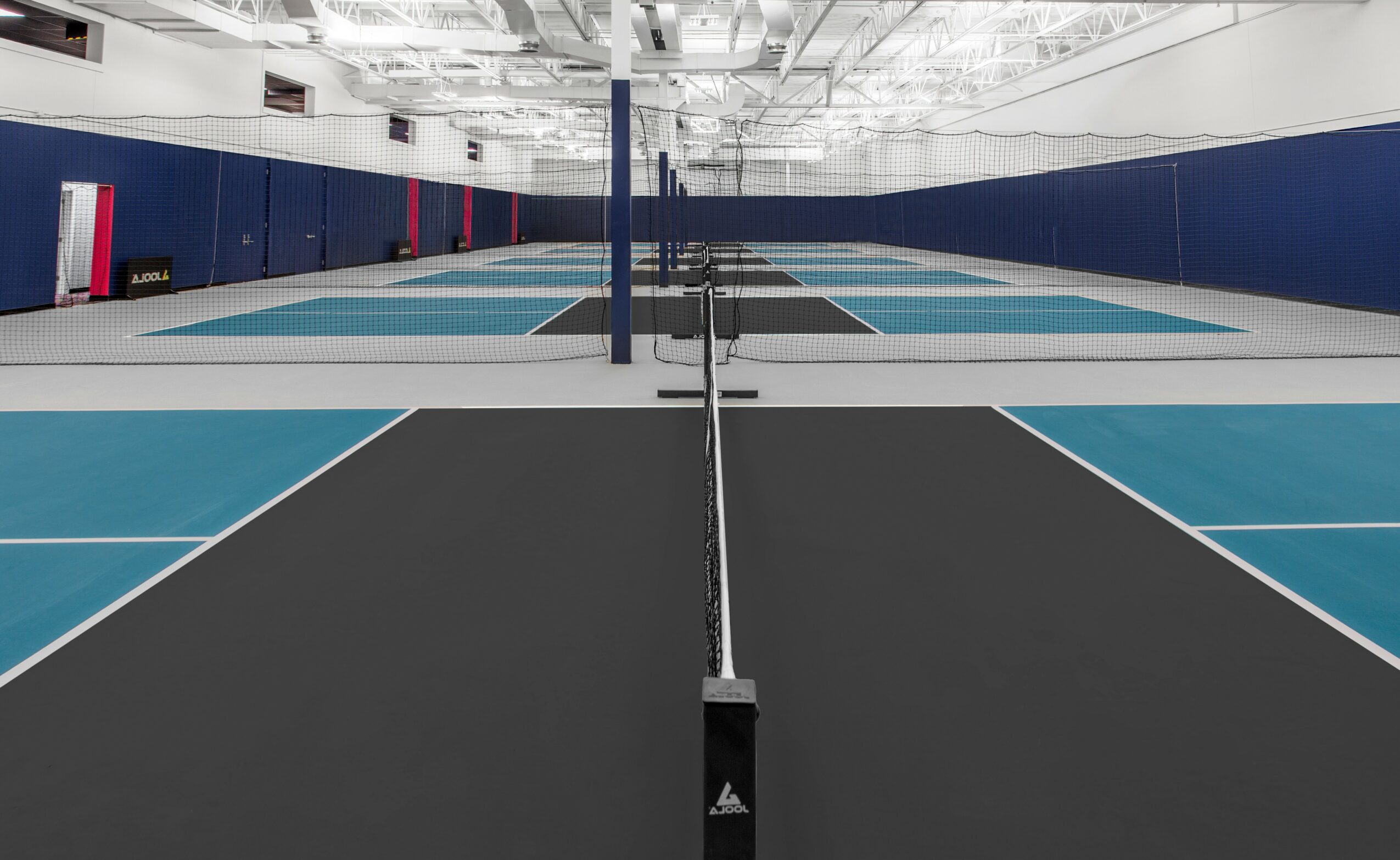 Indoor sports court with blue and black sections, volleyball net in the center, and high ceiling with lights.