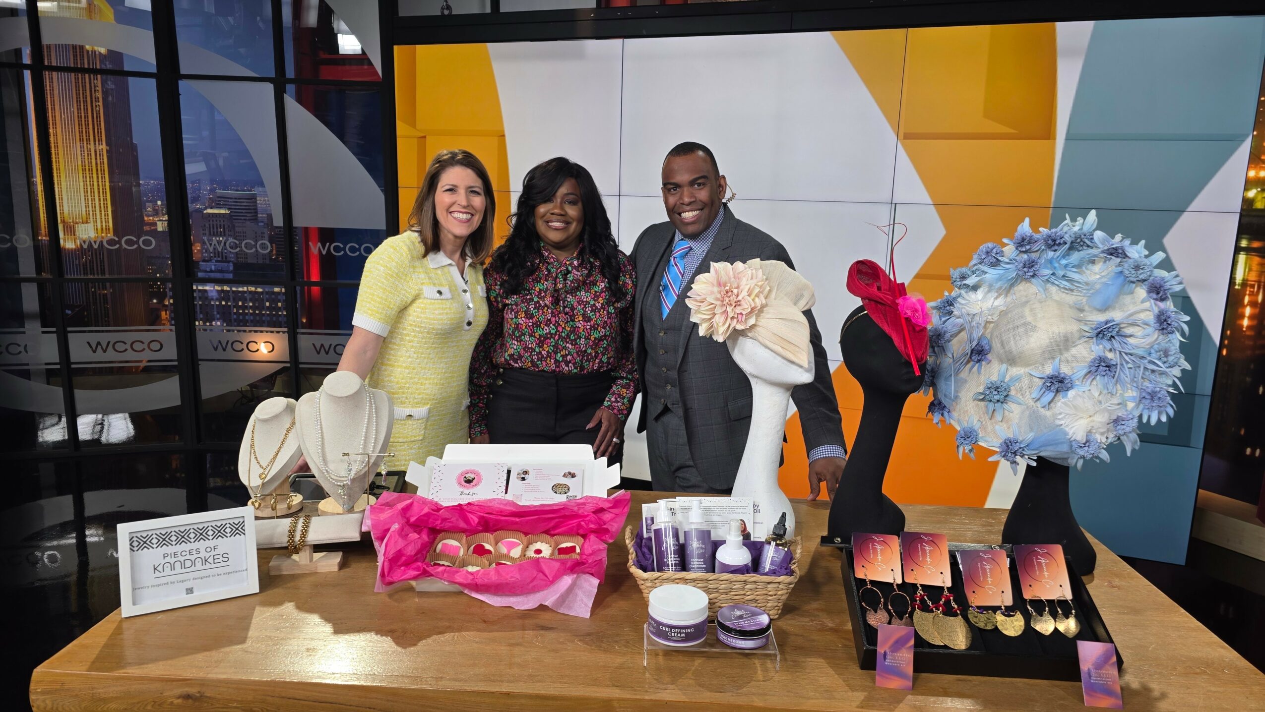 Three people standing behind a table with flowers, gift baskets, and a sign, in a brightly decorated indoor space.