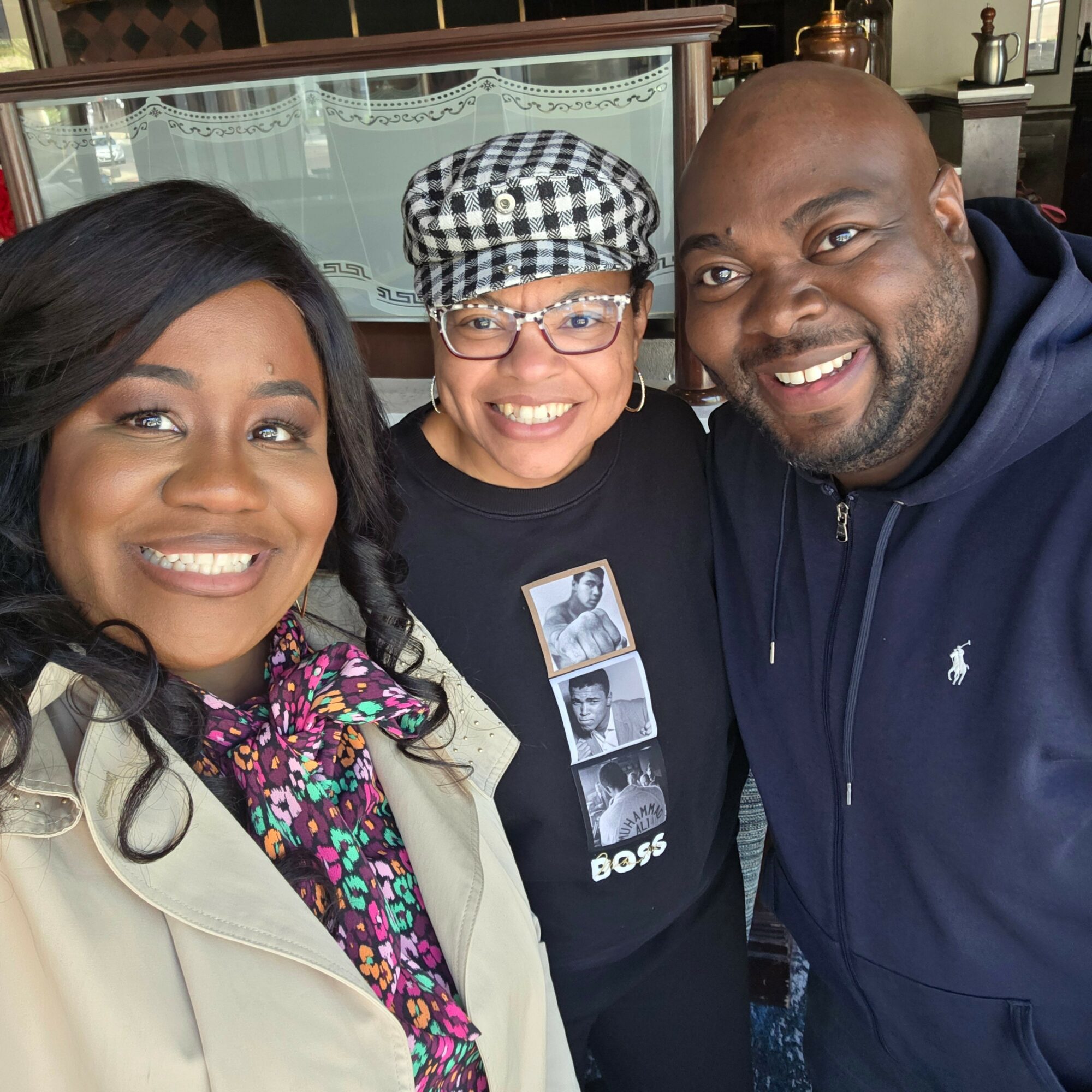 Three people smiling closely for a selfie indoors, two women and one man, with a glass display case in the background.