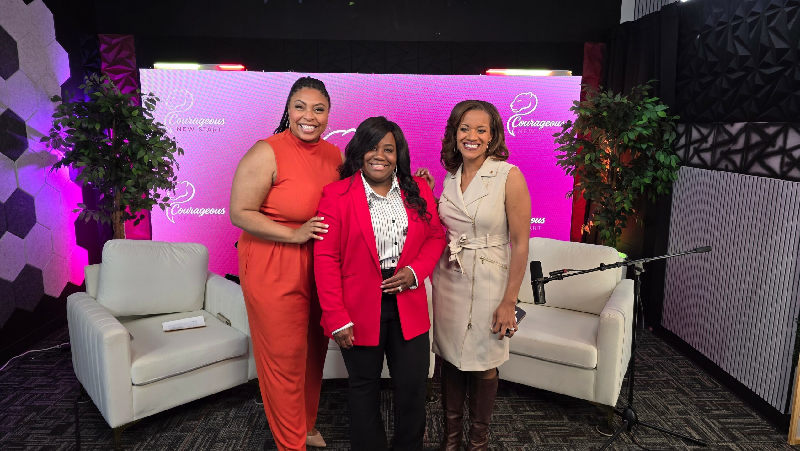 Three women standing in front of a pink backdrop with two white chairs and potted plants, smiling.