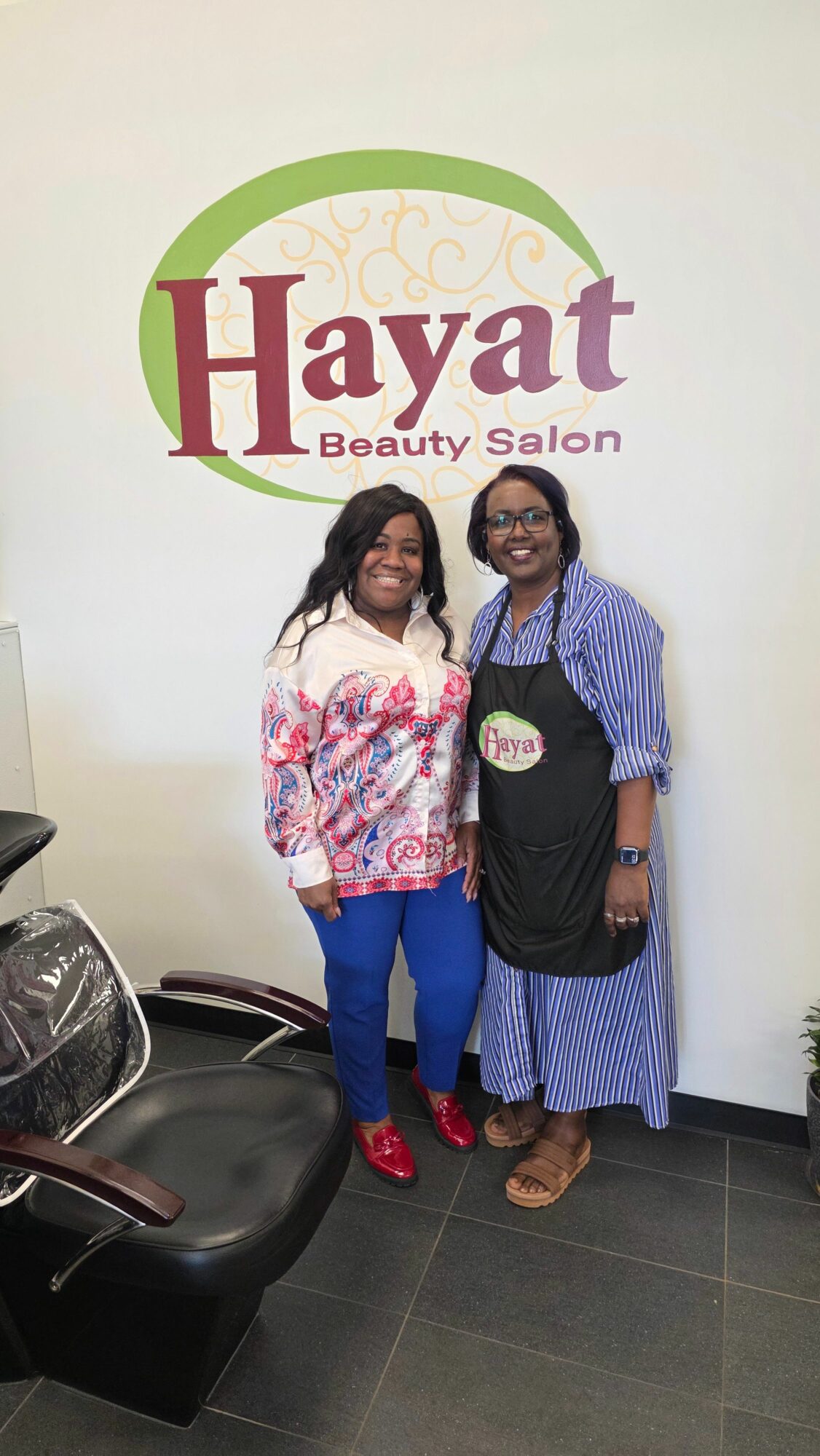 Two women standing in front of a wall with 'Hayat beauty salon' logo, smiling, one wearing a black top and blue skirt, the other in a floral blouse and blue pants.
