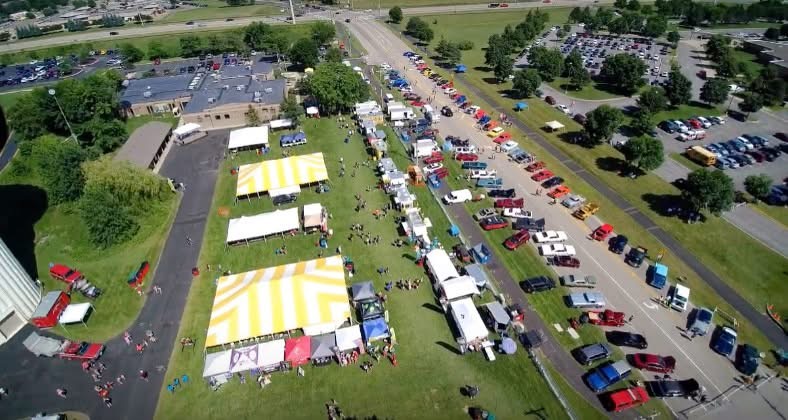 Aerial view of an outdoor event with tents, people, and parked cars on a grassy area and parking lot.
