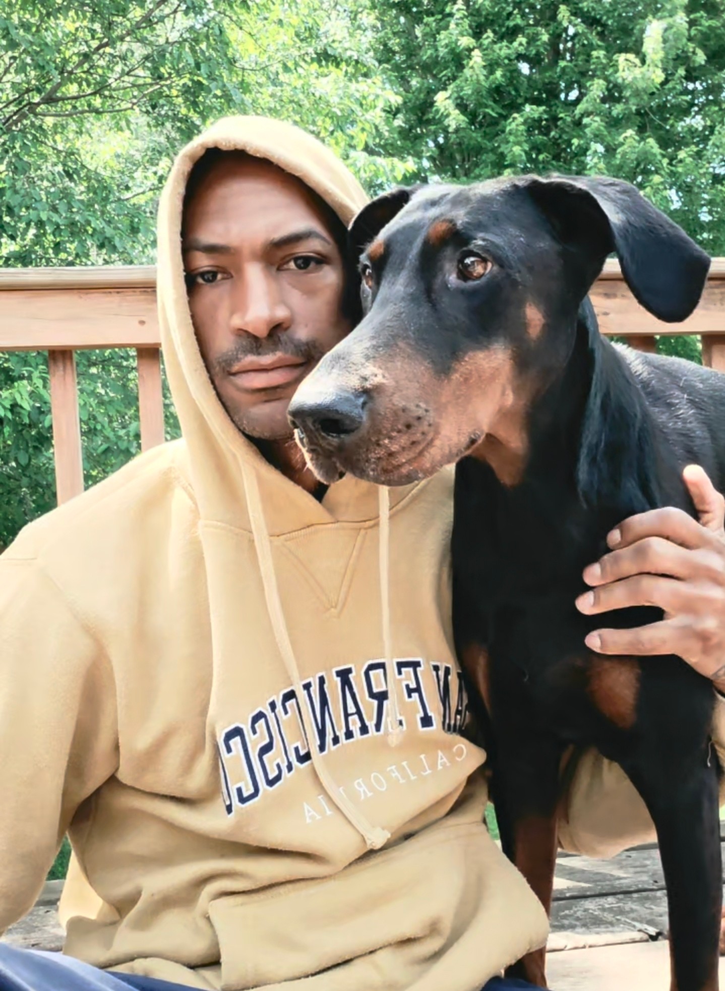 Person in a beige hoodie holding a black and tan dog outdoors with green trees in the background.