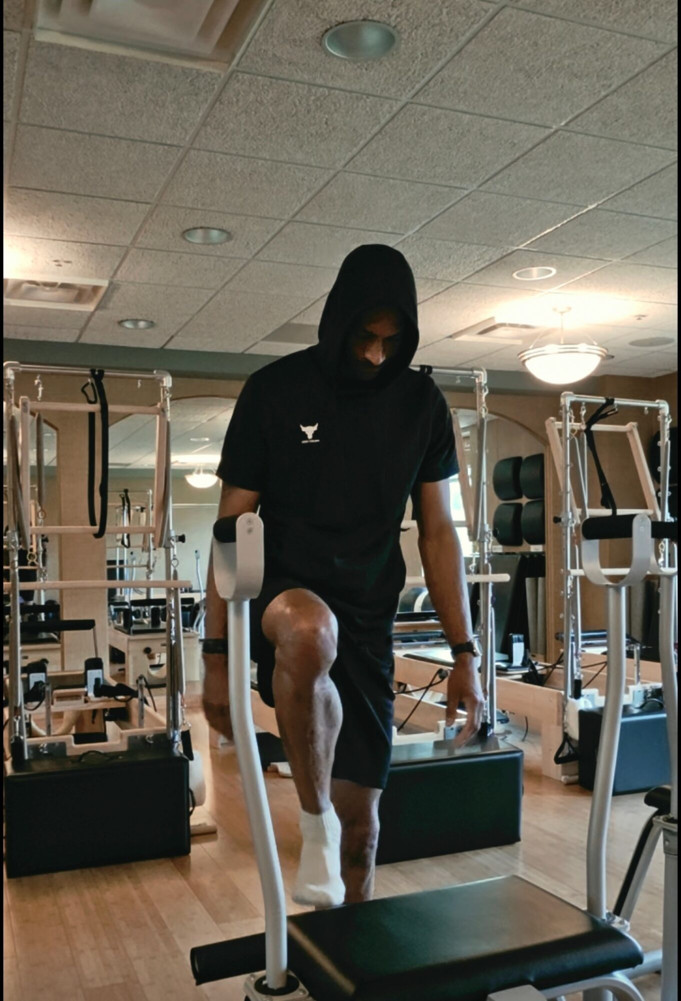 Person with hooded sweatshirt and white pants stepping onto a weight bench in a gym, holding a dumbbell in each hand.