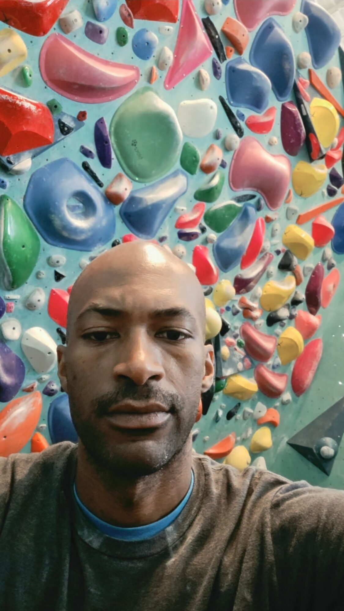 Person taking a selfie in front of a colorful indoor climbing wall with various holds.