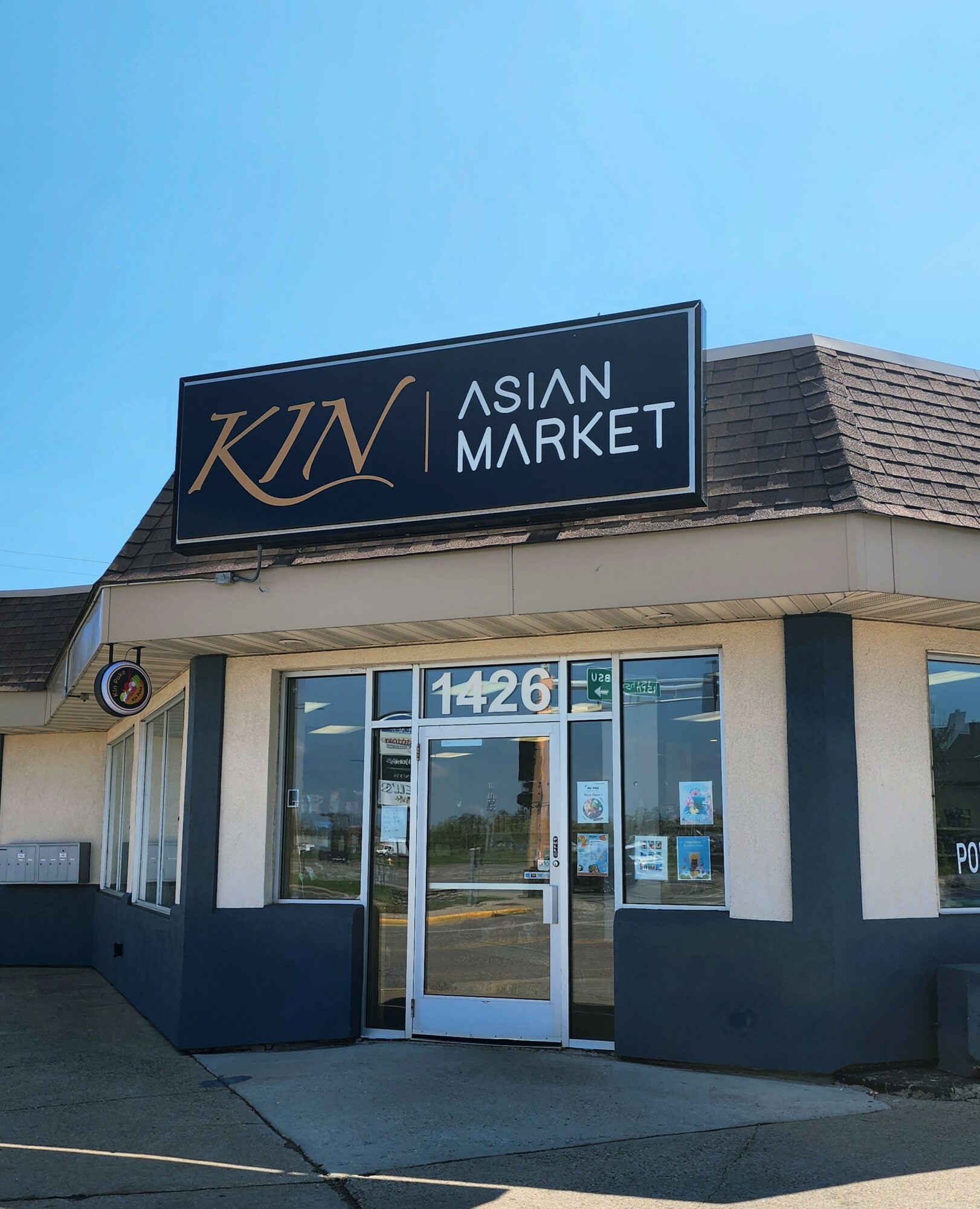Storefront with sign reading 'KIN Asian Market' above entrance, glass doors, and windows, under a blue sky.