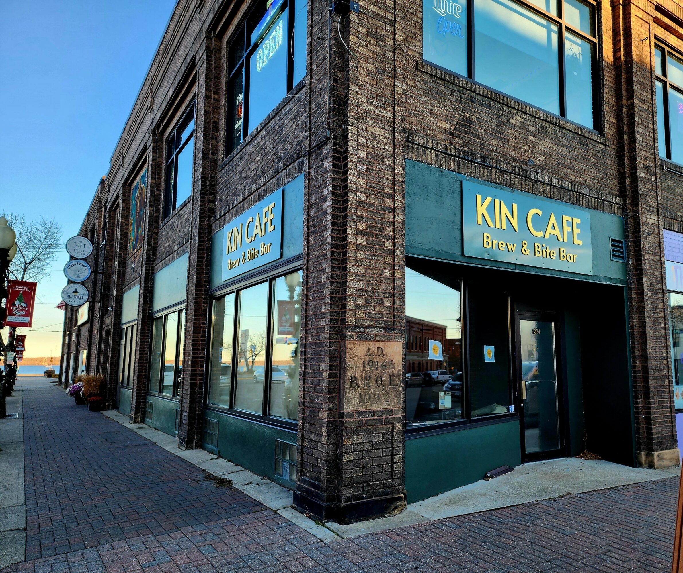 Corner of a brick building with large windows and a blue sign reading 'KIN CAFE Brew & Bk R' on a city sidewalk.