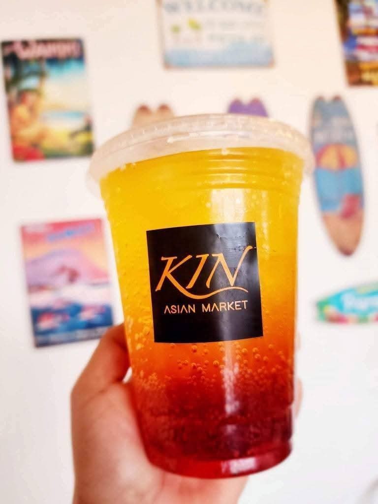 Hand holding a colorful drink with a black label reading 'KIN ASIAN MARKET' against a wall with posters.