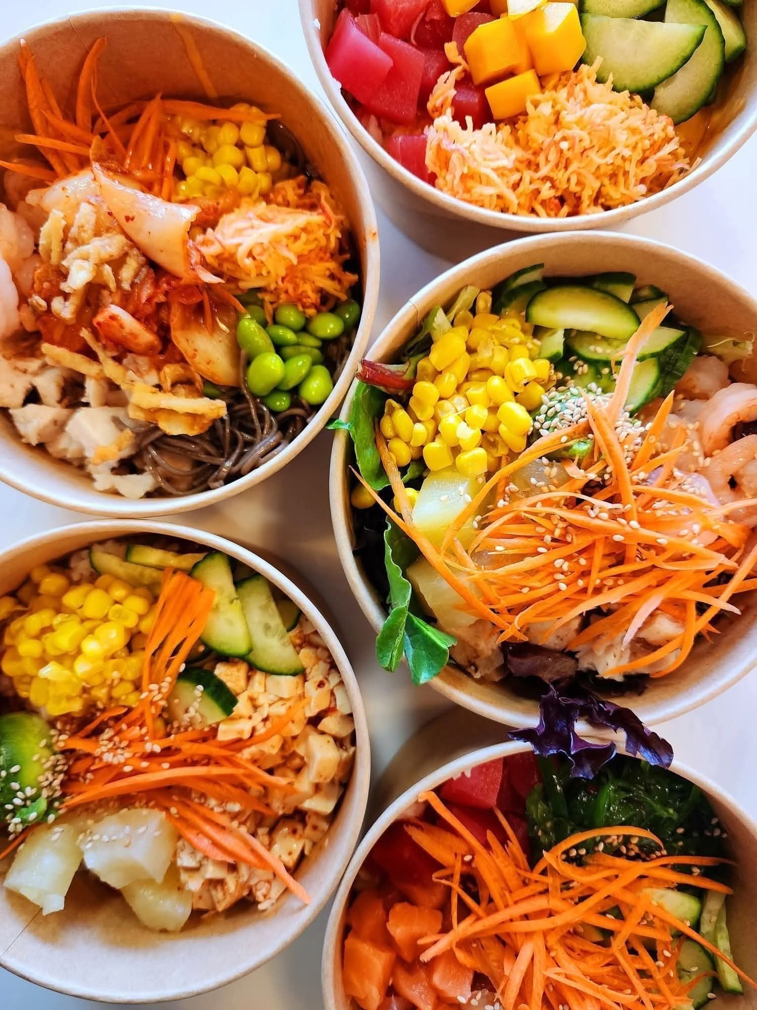 Five bowls of colorful salads with vegetables, shredded carrots, corn, and greens, arranged on a white surface.