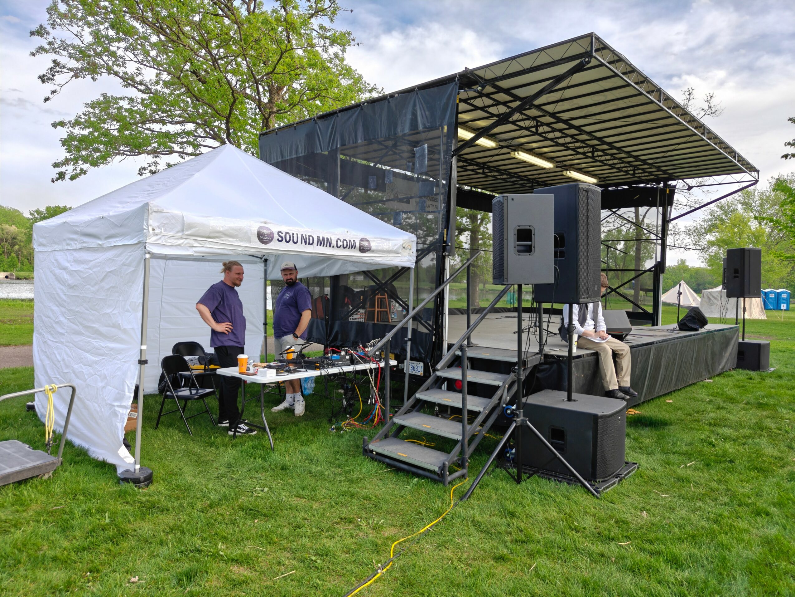 Outdoor stage with sound equipment and a white tent, two people working at a table, green grass, trees, and cloudy sky