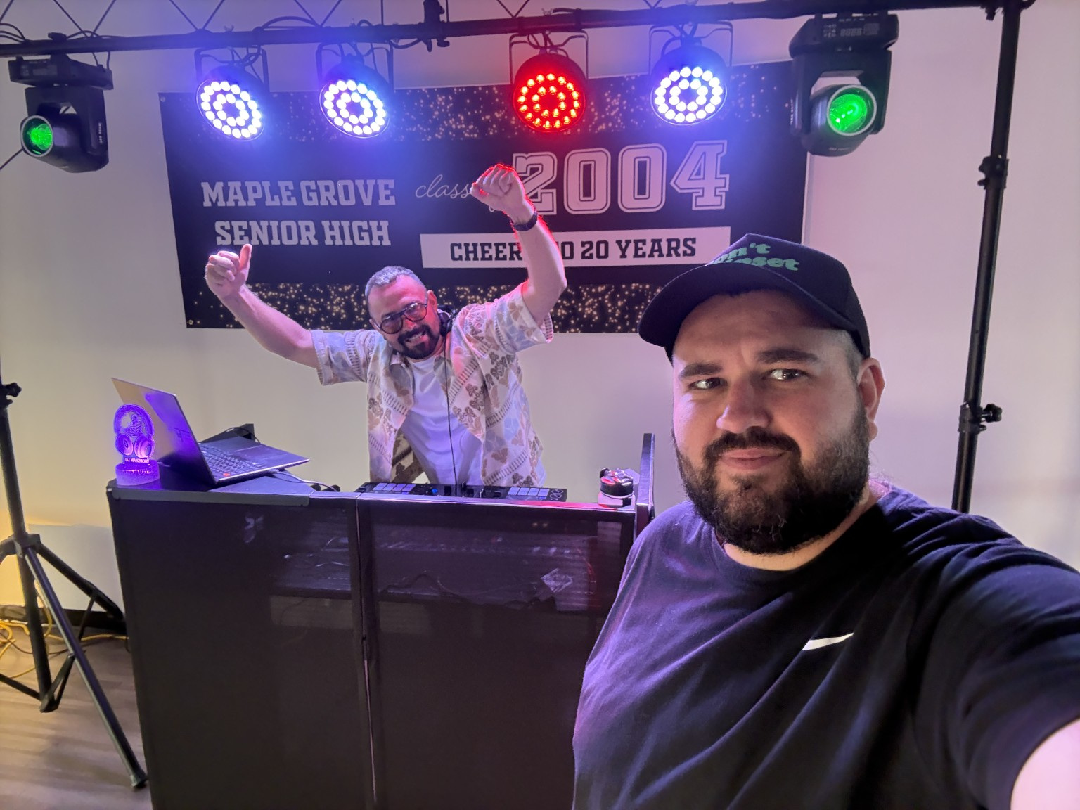 Two men at a celebration event, one taking a selfie and the other cheering behind a DJ booth, with a banner in the background.