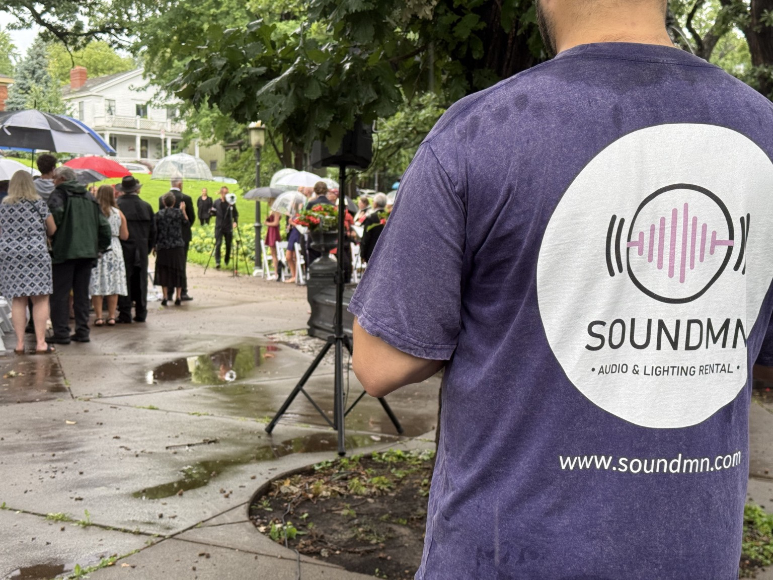 Person wearing a purple SoundMN t-shirt standing outdoors near a group of people with umbrellas, trees, and buildings in the background.