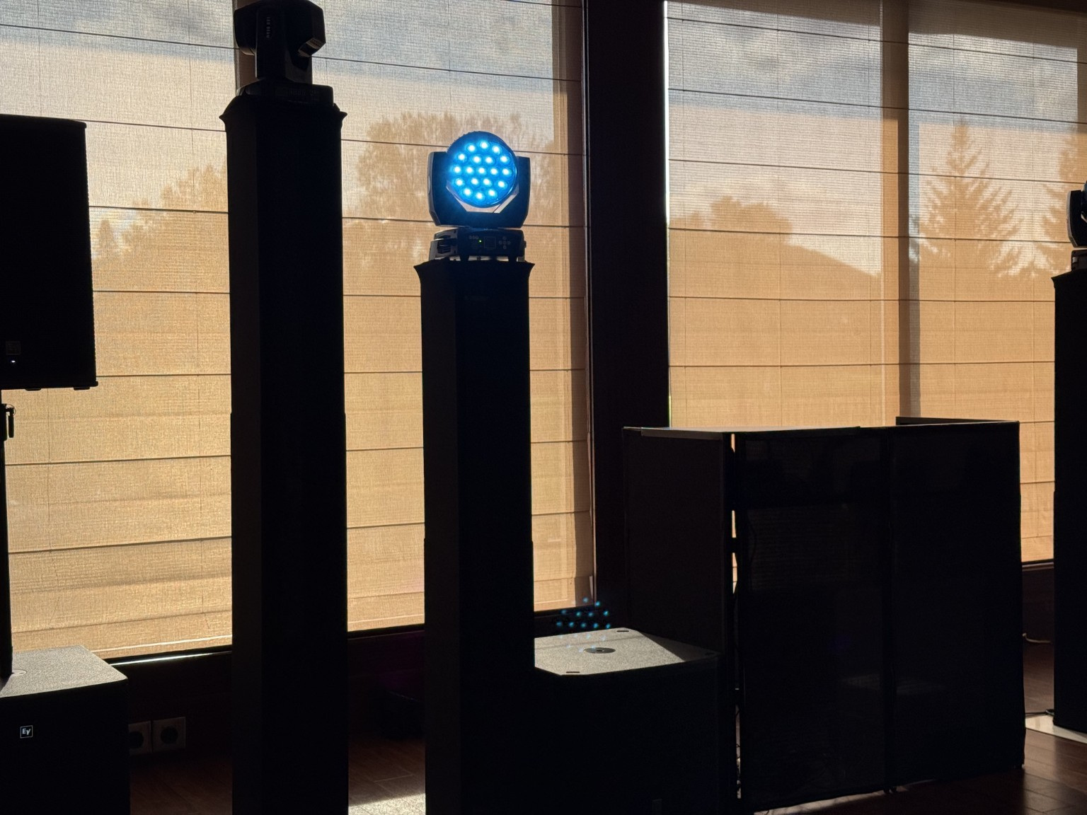 Blue stage light on a black stand in front of a window with blinds, sunlight outside, and equipment nearby.