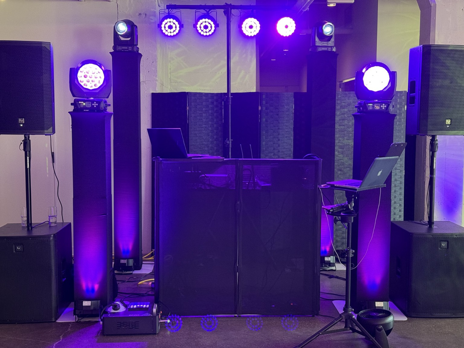 DJ setup with speakers, lights, and equipment in a dimly lit room, illuminated by purple and blue lights.