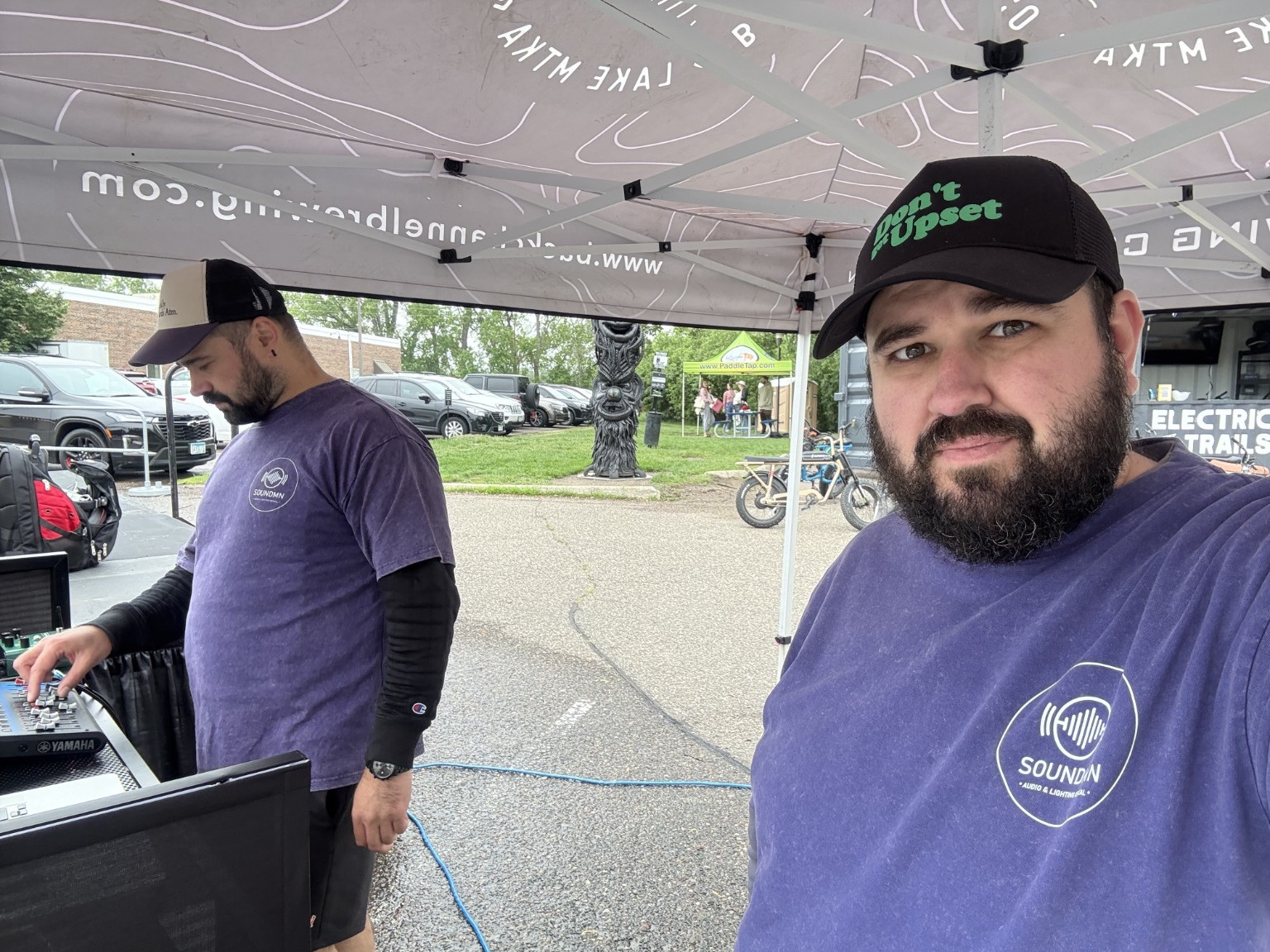 Two men in purple shirts and black caps under a canopy, one looking at a device, the other smiling at the camera.