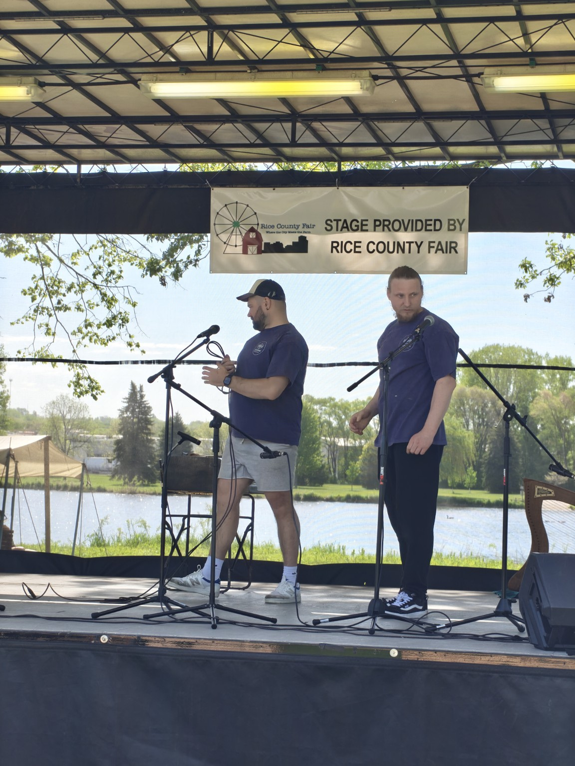 Two men stand on an outdoor stage with microphones, a lake, and trees in the background, under a metal roof.