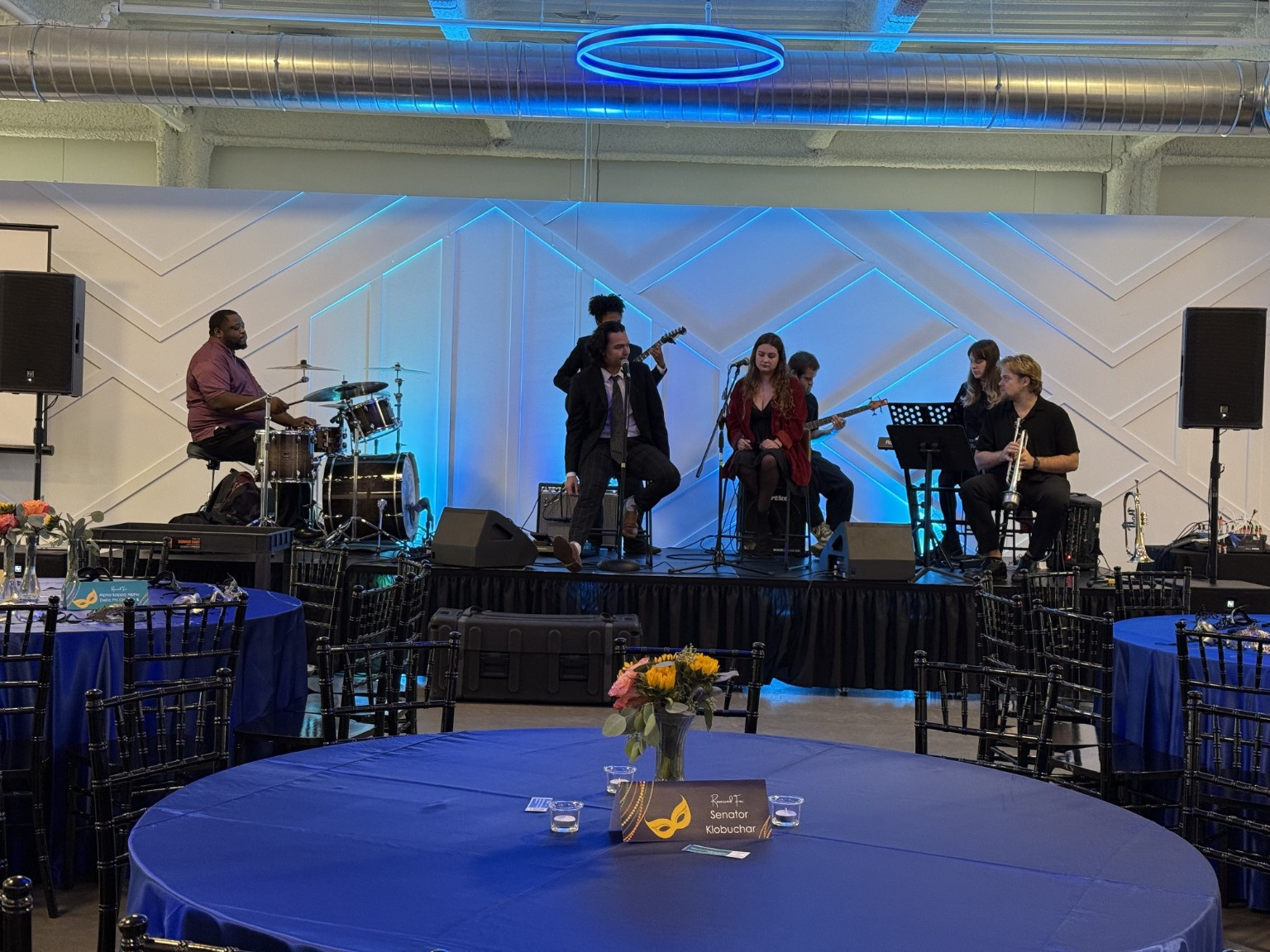 Band performing on stage with four musicians, colorful lighting, and empty tables with blue tablecloths in foreground.