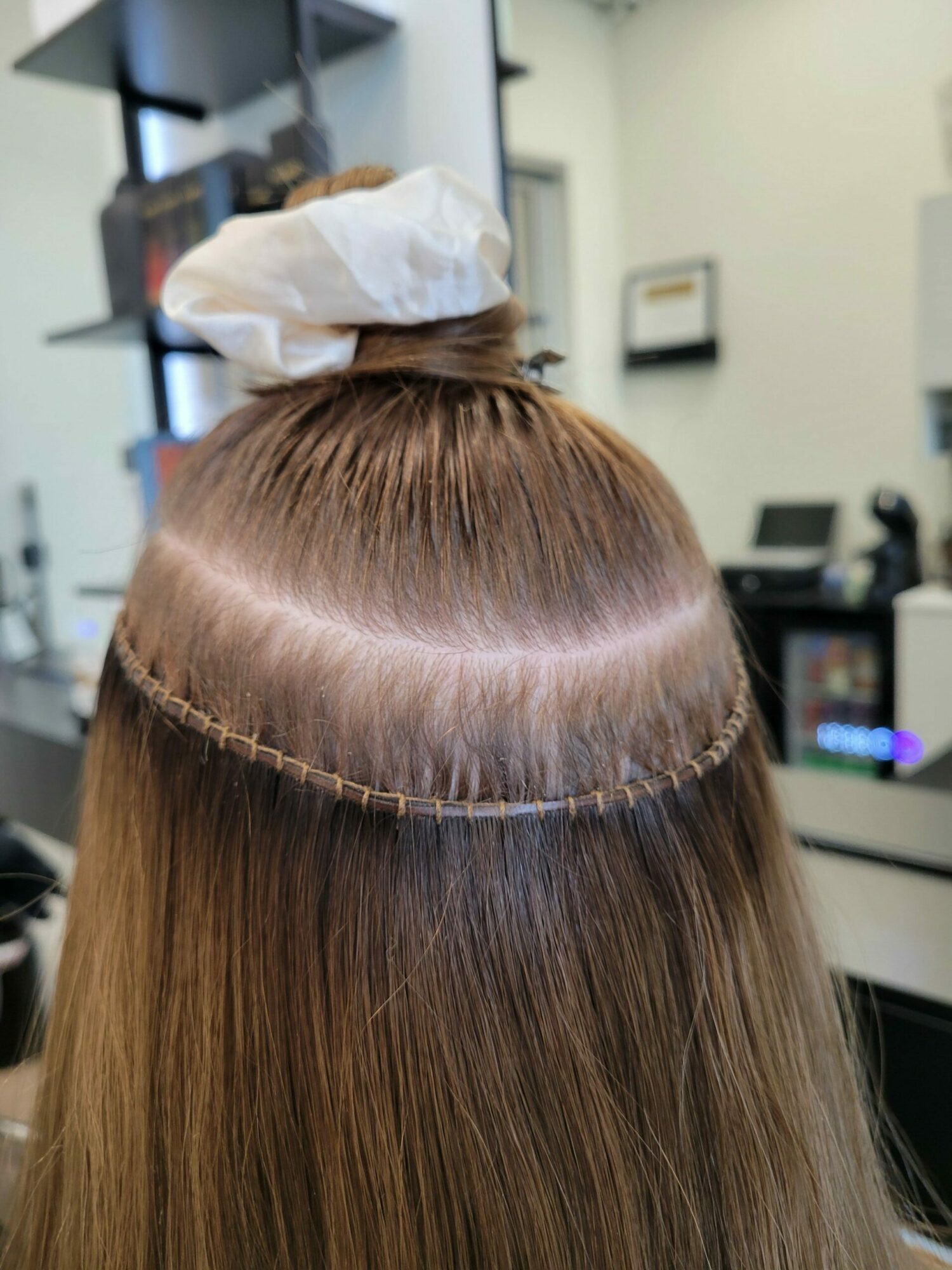 Back of a person's head with long, straight hair tied with a white bow, in a salon setting.