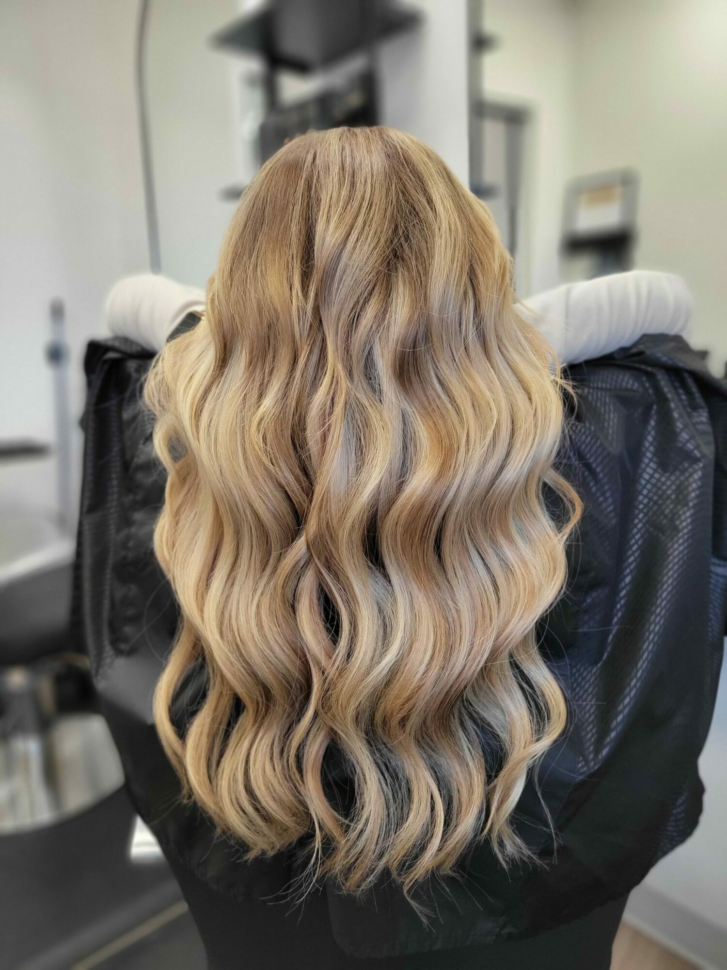 Person with long, wavy blonde hair sitting in a salon chair, facing away from the camera.