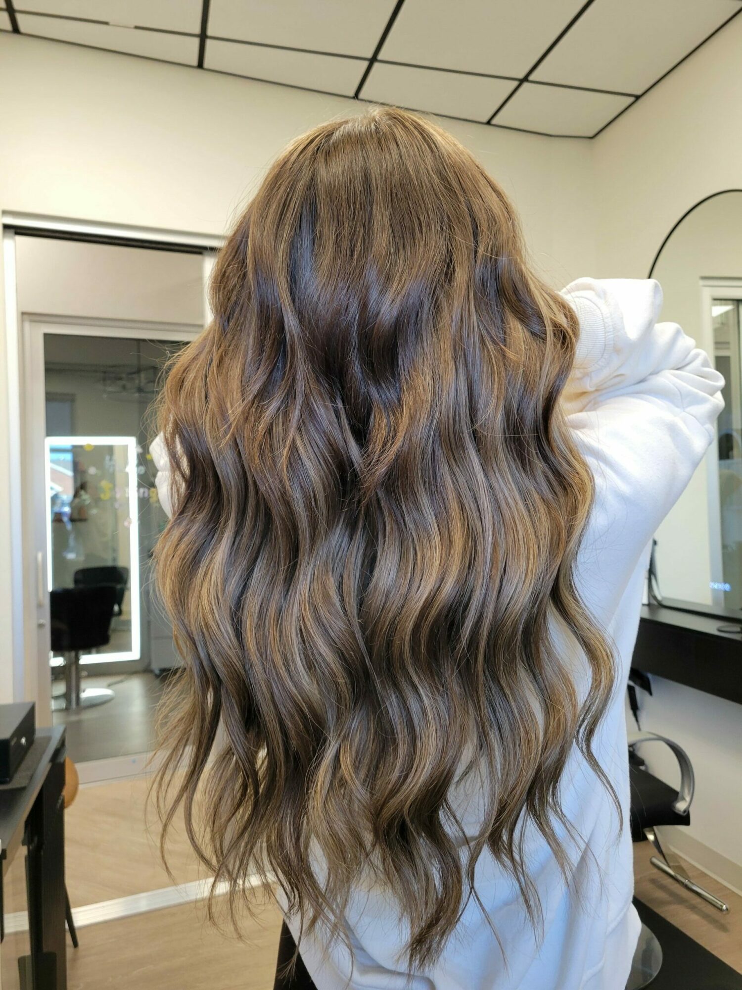 Person with long, wavy hair standing in a room with a window and tiled wall, facing away from the camera.