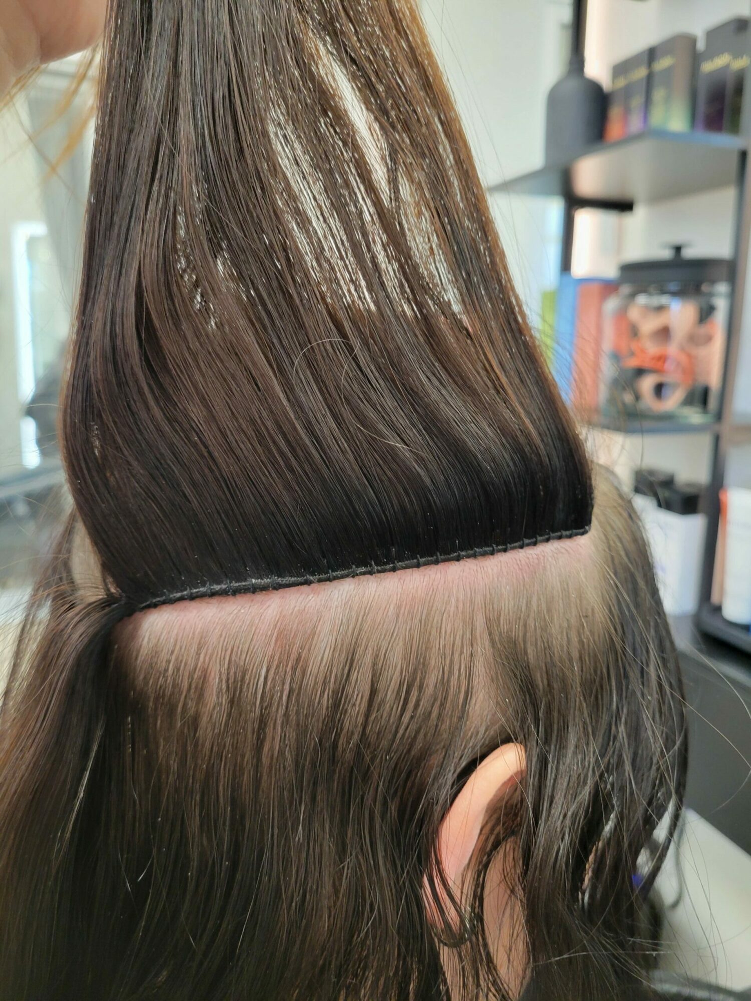 Close-up of a person's head showing long, straight, dark brown hair parted to the side, with a background of bookshelves.
