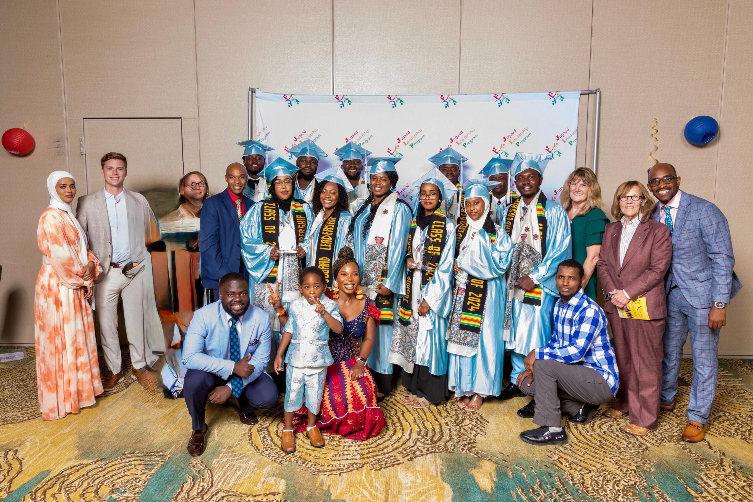 Group of people, including children and adults, in graduation gowns and caps, posing indoors with a decorated backdrop.