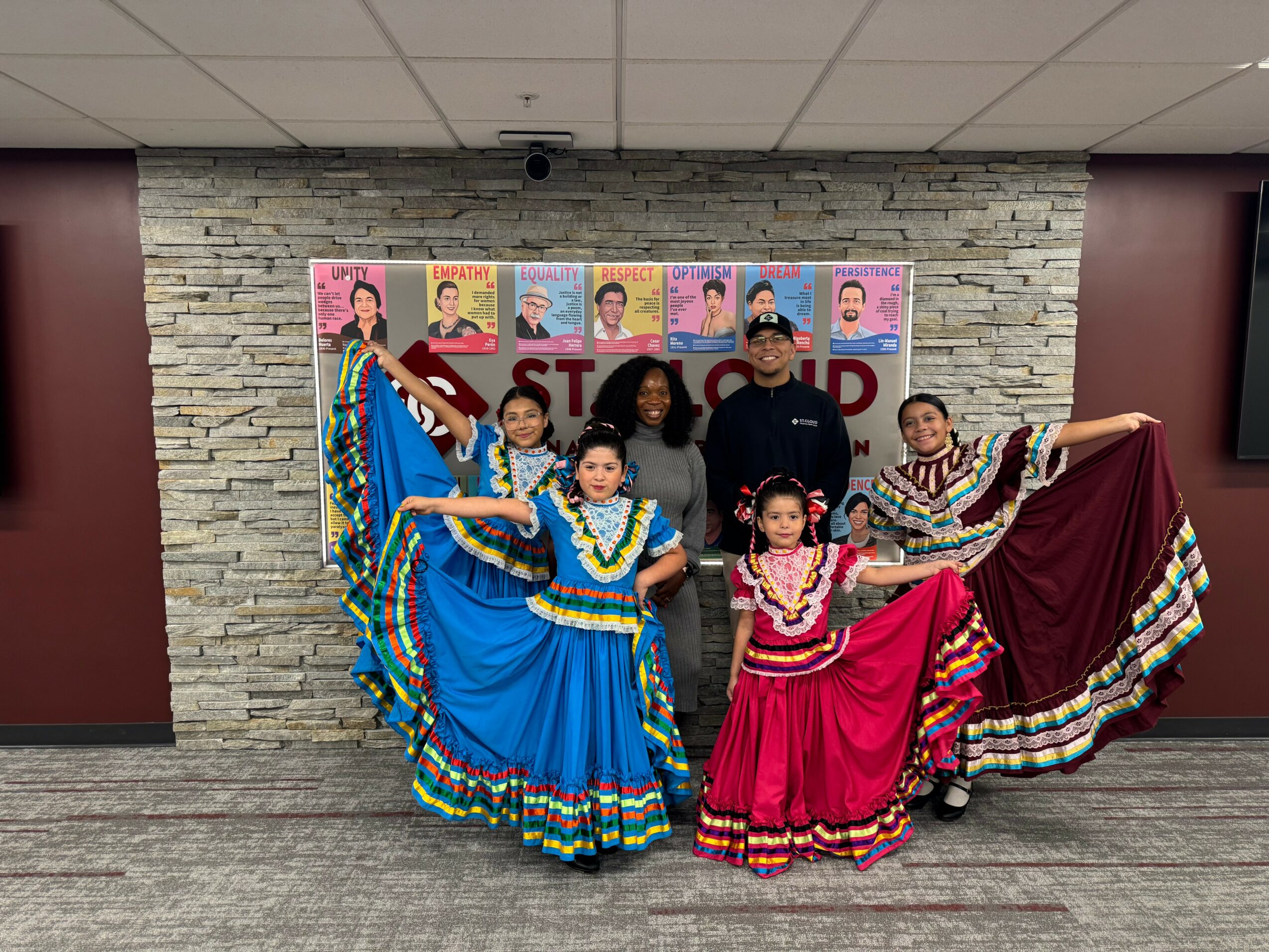 Group of children and adults in colorful traditional dresses posing indoors with a stone wall background and posters, some holding dresses.