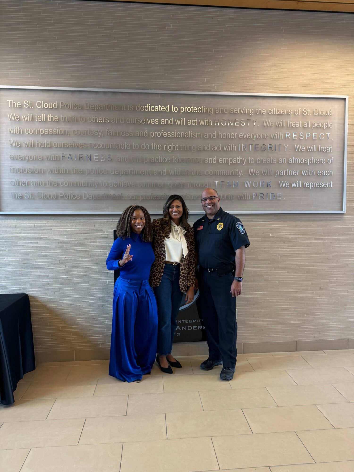 Three women standing together in front of a wall with a plaque, smiling, one in a blue dress, one in a white top and leopard print jacket, and one in a police uniform.