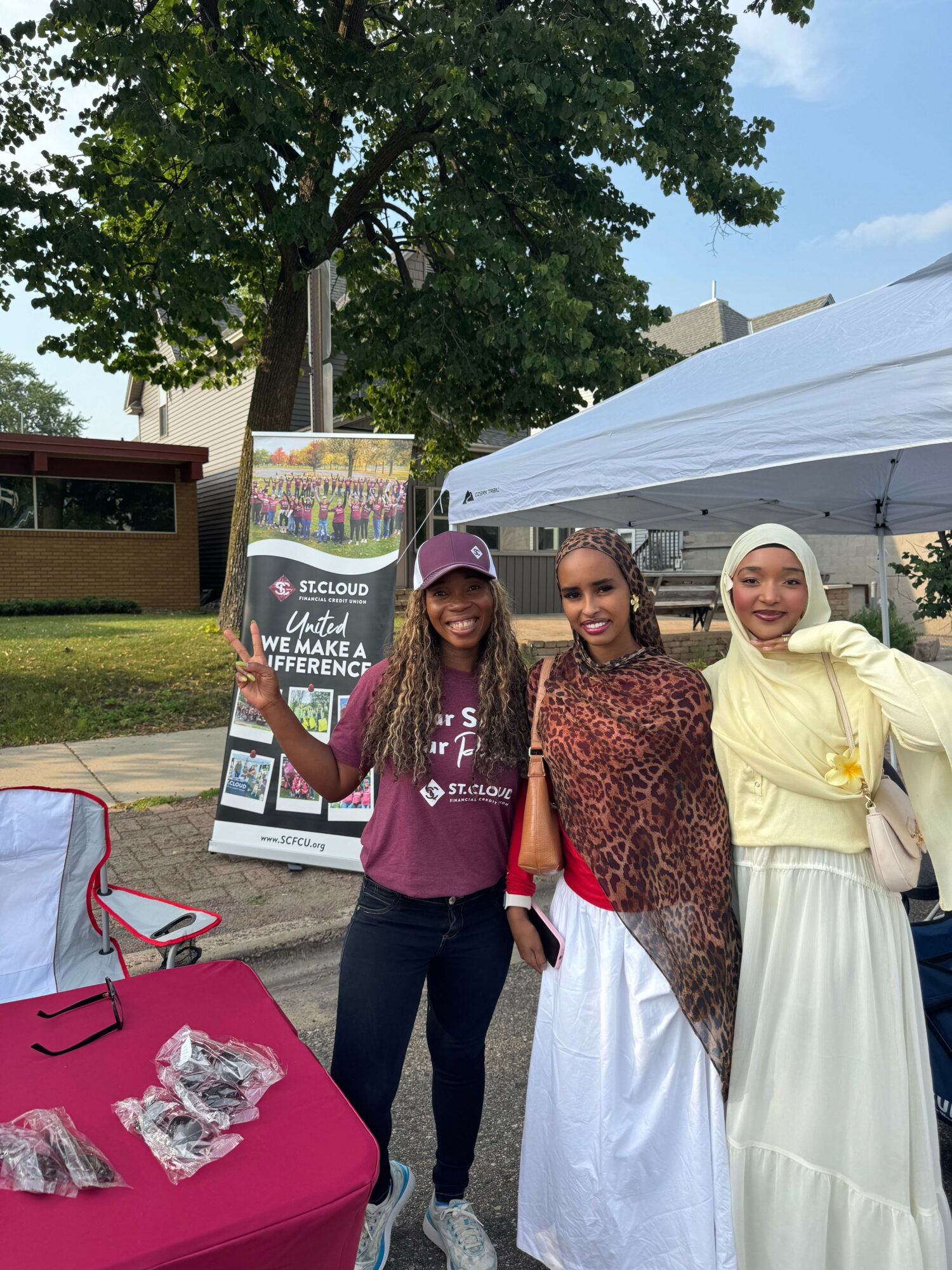 Three women standing outdoors near a tent and a signboard, with trees and a building in the background.