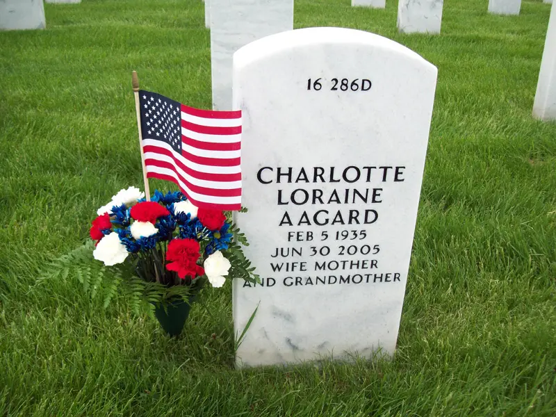Gravestone with American flag and flowers, inscribed with name, birth and death dates, and titles, in a cemetery.