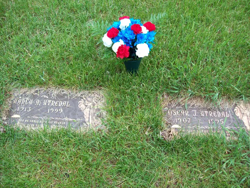 Two grave markers with flowers on grass, one with a bouquet of red, white, and blue flowers, both graves marked with dates.