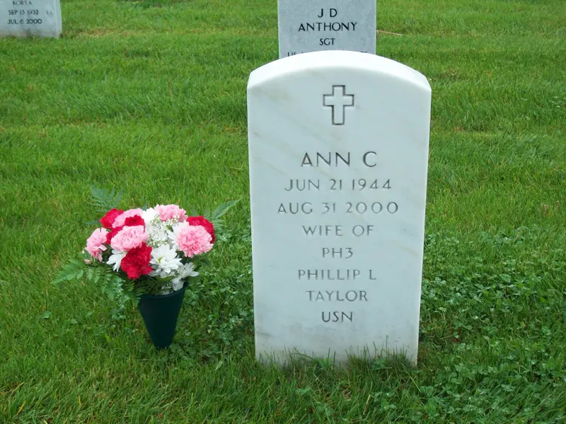 White gravestone with a cross, inscription for Ann C., with a bouquet of pink and white flowers nearby, on green grass.