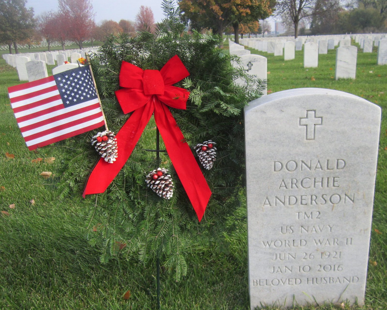 Gravestone with a wreath and American flag at a cemetery, autumn trees in background.