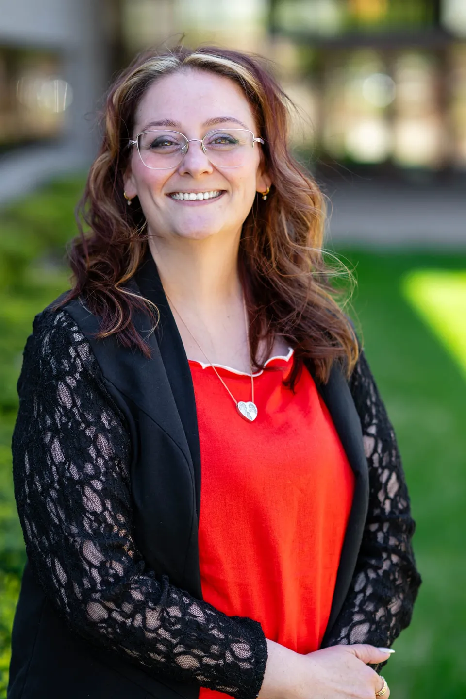 Smiling woman with glasses and wavy hair outdoors, wearing a red top and black lace cardigan.