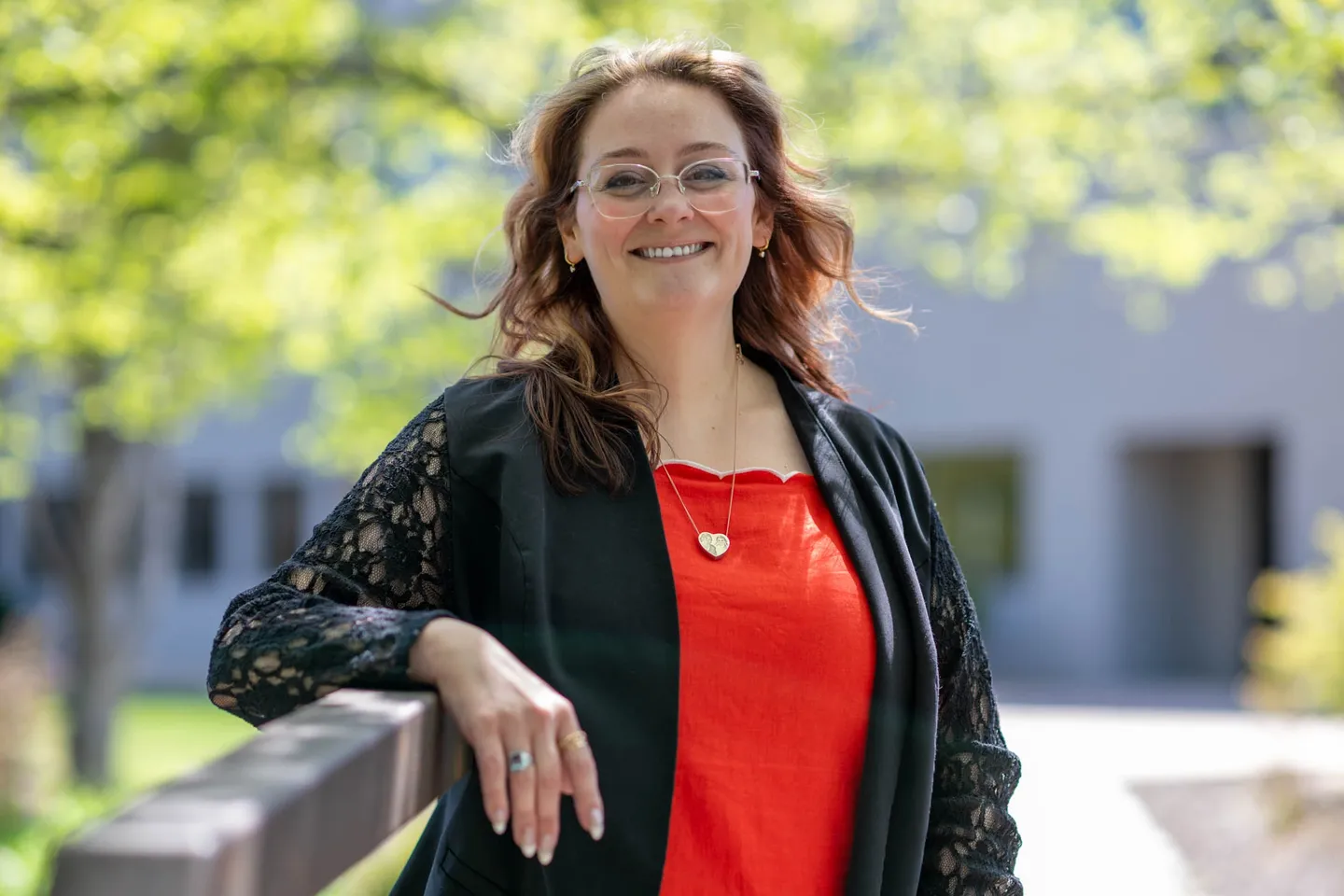 Smiling woman with glasses and wavy hair outdoors, wearing a red top and black lace cardigan, leaning on a railing.